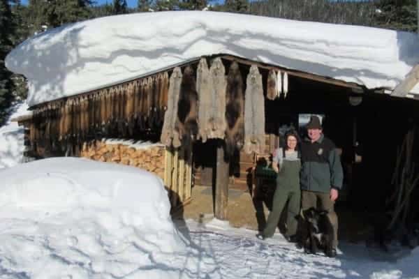 A man and a woman stand outside a log cabin in winter with furs from a season of trapping hanging on the wall