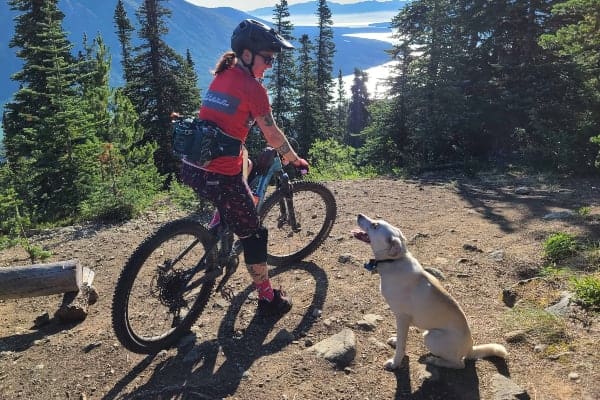 A woman on a mountain bike looking down at a dog who is looking back up at her