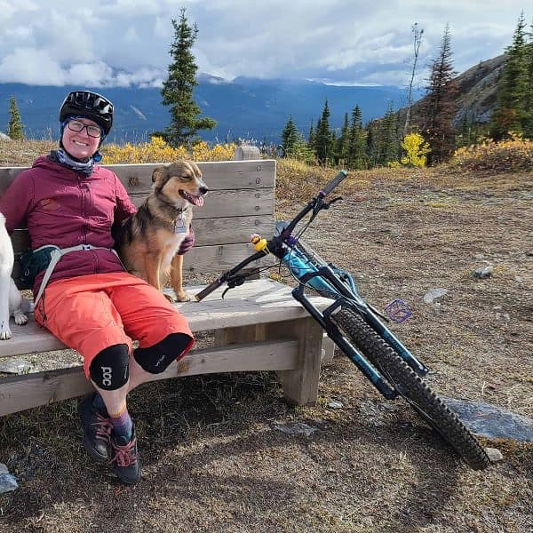 A woman and two dogs sit on a bech with a mountain bike nearby